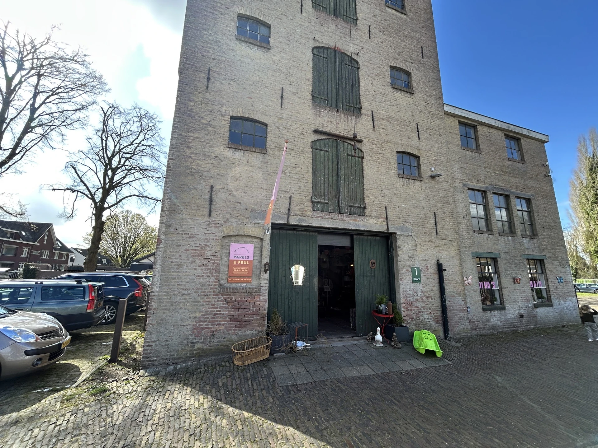 Bakstenen gevel van kringloopwinkel met groene deuren en ramen, parkeerplaats met auto's ervoor