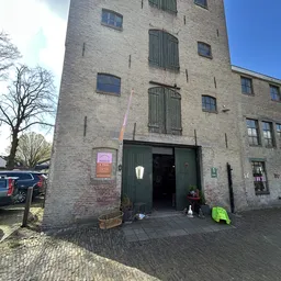 Bakstenen gevel van kringloopwinkel met groene deuren en ramen, parkeerplaats met auto's ervoor