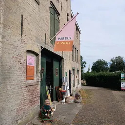 Bakstenen gevel van kringloopwinkel met groene deur, oranje-roze uithangbord met 'PARELS & PRUL', spullen voor de ingang