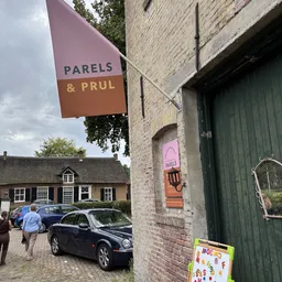 Voorgevel van kringloopwinkel met bruin-oranje uithangbord 'PARELS & PRUL' en groene deur, bakstenen gevel
