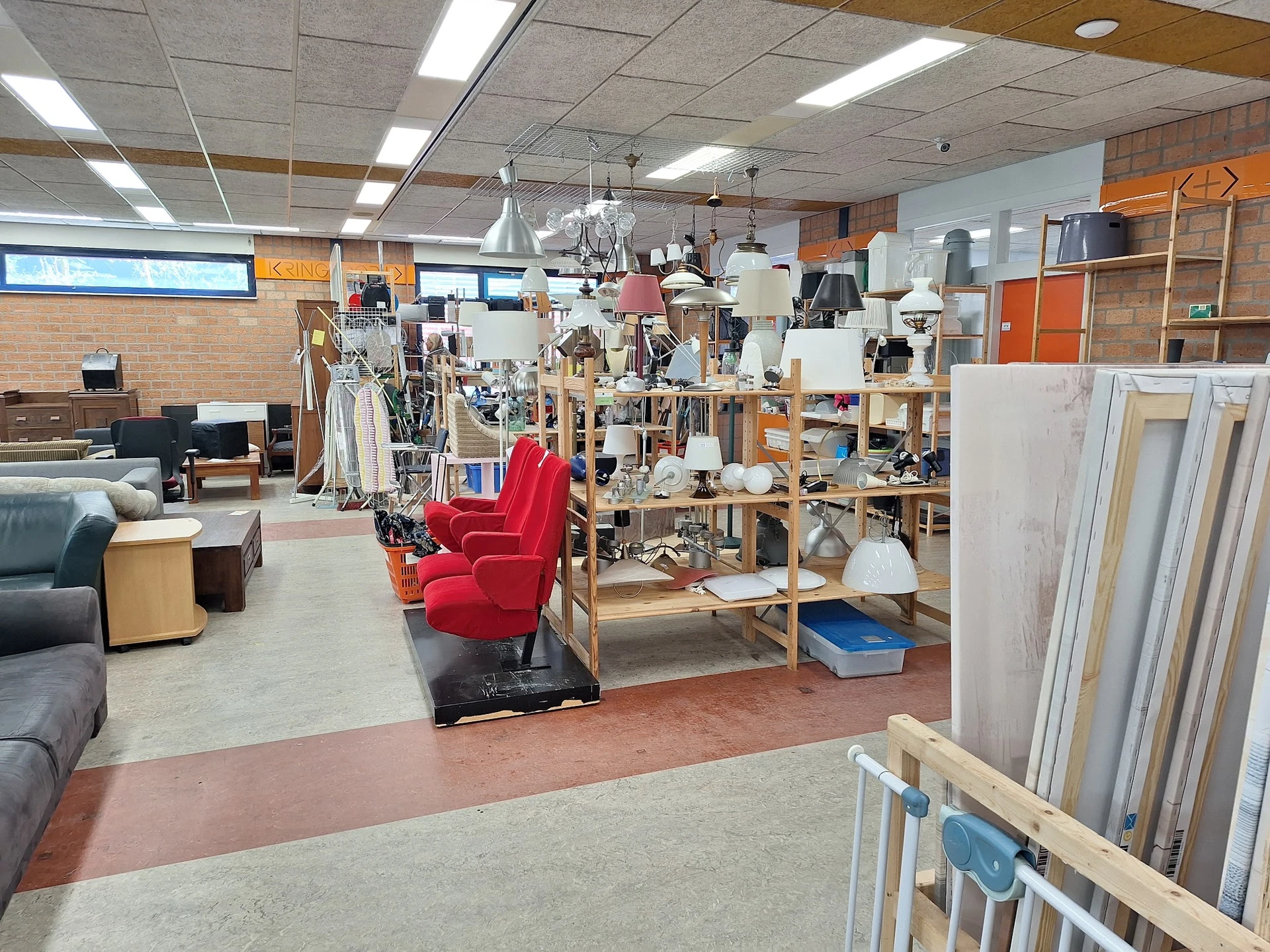 Winkelvloer van kringloopwinkel met stellingen vol spullen, rode fauteuil, kleding aan rekken en diversen huishoudartikelen