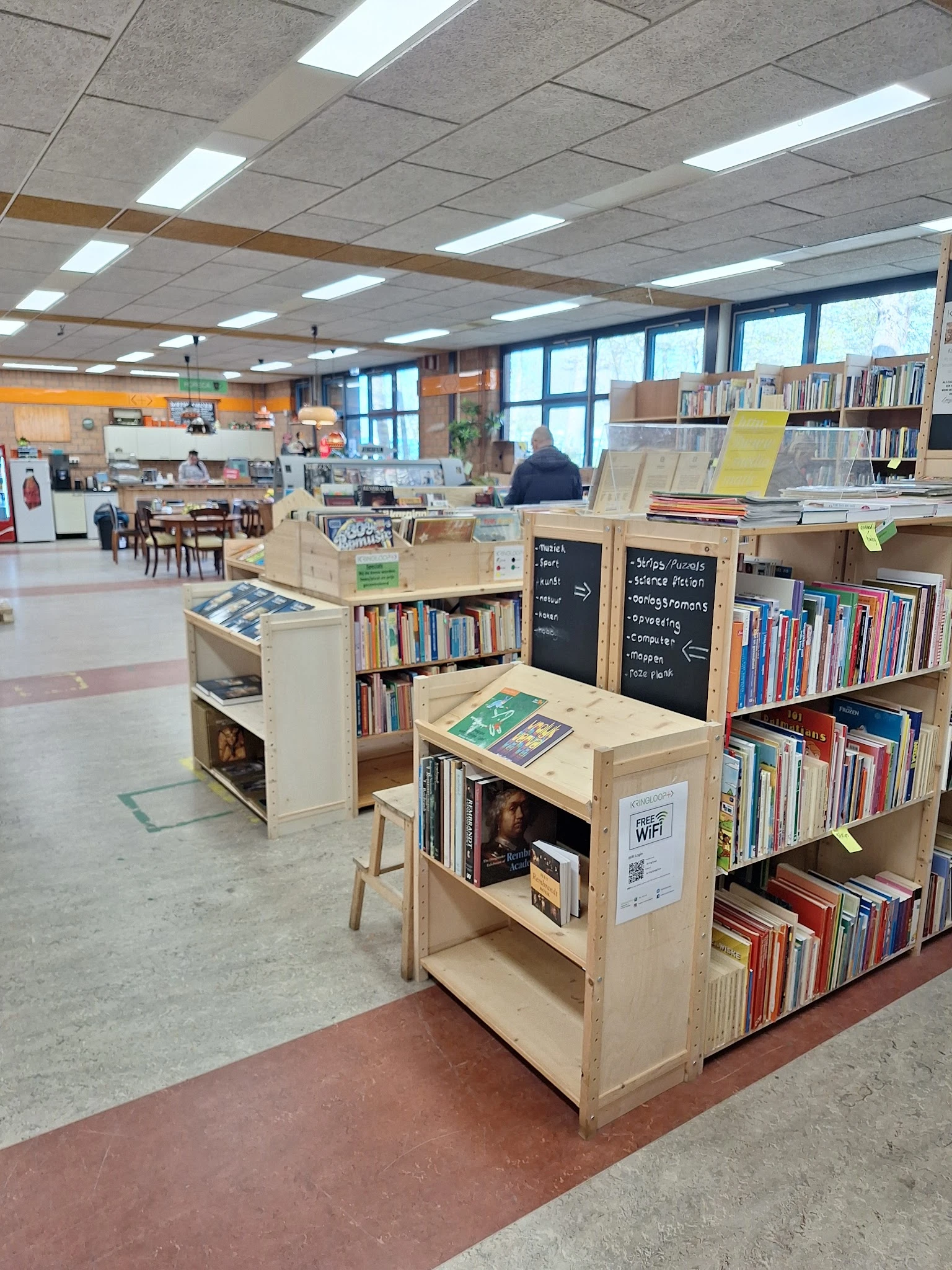 Winkelvloer met boekenkasten, stellingen met boeken en media, leestafel met gele blad, ramen met daglicht