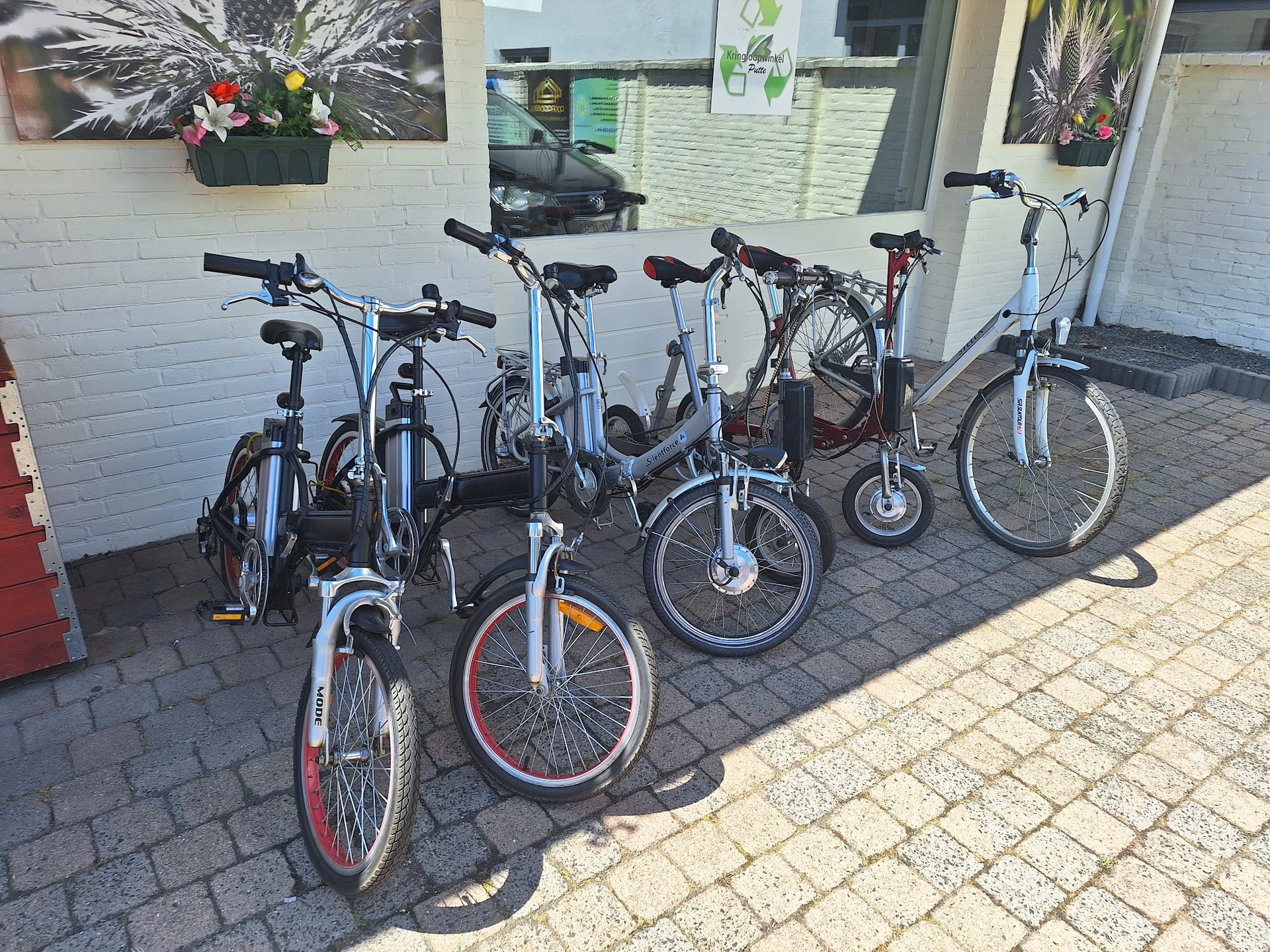 Rij van verschillende tweedehands fietsen en e-bikes buiten tegen een witte muur