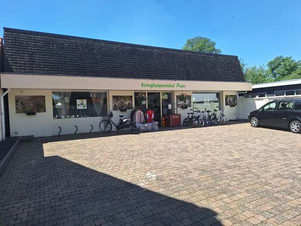 Voorkant van kringloopwinkel met witte pui, donker dak en parkeerplaats met enkele auto's