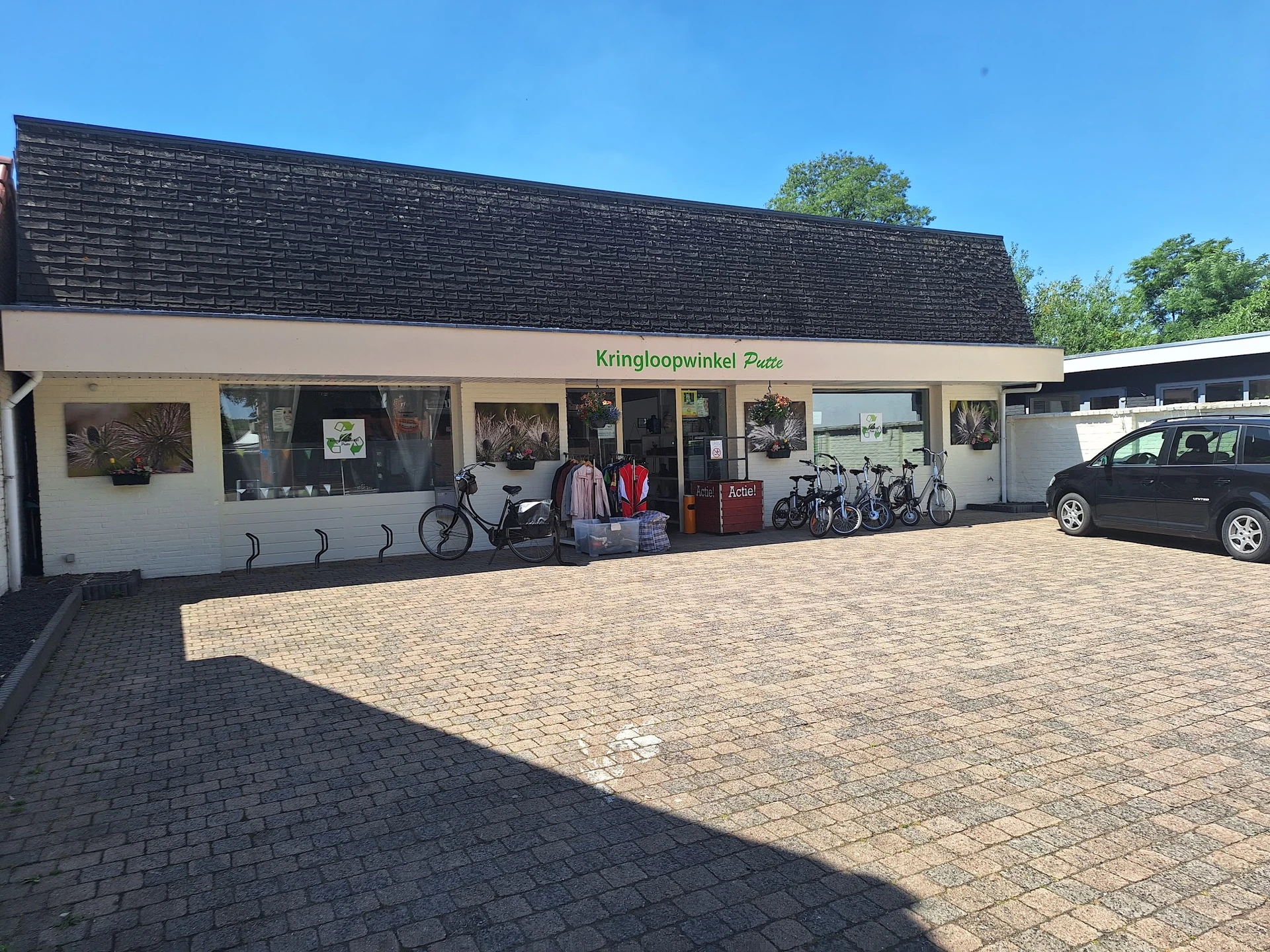 Voorkant van kringloopwinkel met witte pui, donker dak en parkeerplaats met enkele auto's