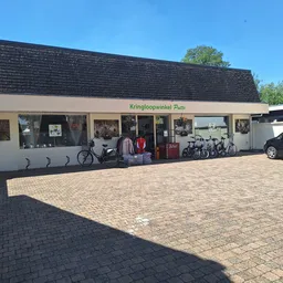 Voorkant van kringloopwinkel met witte pui, donker dak en parkeerplaats met enkele auto's