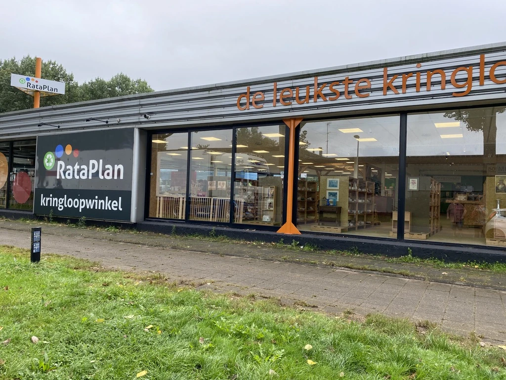 Voorkant van kringloopwinkel met grote ramen, donkerblauwe gevel en oranje accenten, zicht op interieur met stellingen