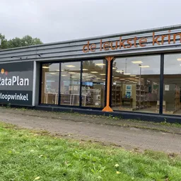 Voorkant van kringloopwinkel met grote ramen, donkerblauwe gevel en oranje accenten, zicht op interieur met stellingen