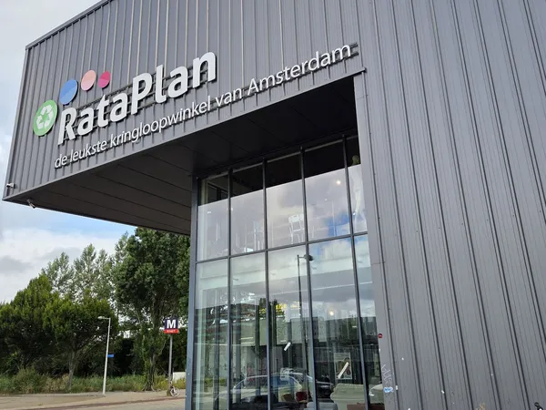 Grijze gevel van kringloopwinkel RataPlan met uitstekend logo en glazen ingang, omgeven door groen
