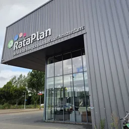 Grijze gevel van kringloopwinkel RataPlan met uitstekend logo en glazen ingang, omgeven door groen