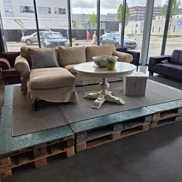Winkelinterieur met meubels: beige fauteuils, zwarte bank en wit tafeltje op grijs tapijt, grote ramen met uitzicht op straat