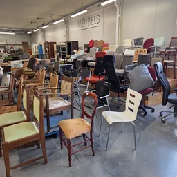 Verschillende meubels en stoelen uitgestald in een kringloopwinkel, waaronder houten stoelen, ergonomische bureaukstoelen en 