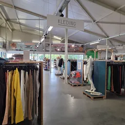 Winkelvloer met kledingrekken vol tweedehands kleding, hoog plafond met lichtkappen, bordje 'KLEDING' zichtbaar