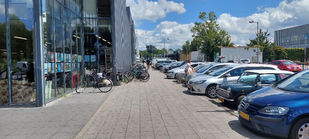 Voorgevel van kringloopwinkel met fietsen geparkeerd links en parkeerplaats met auto's rechts
