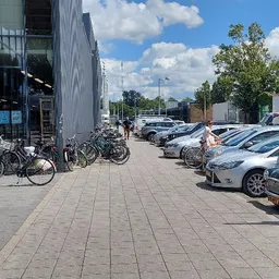 Voorgevel van kringloopwinkel met fietsen geparkeerd links en parkeerplaats met auto's rechts
