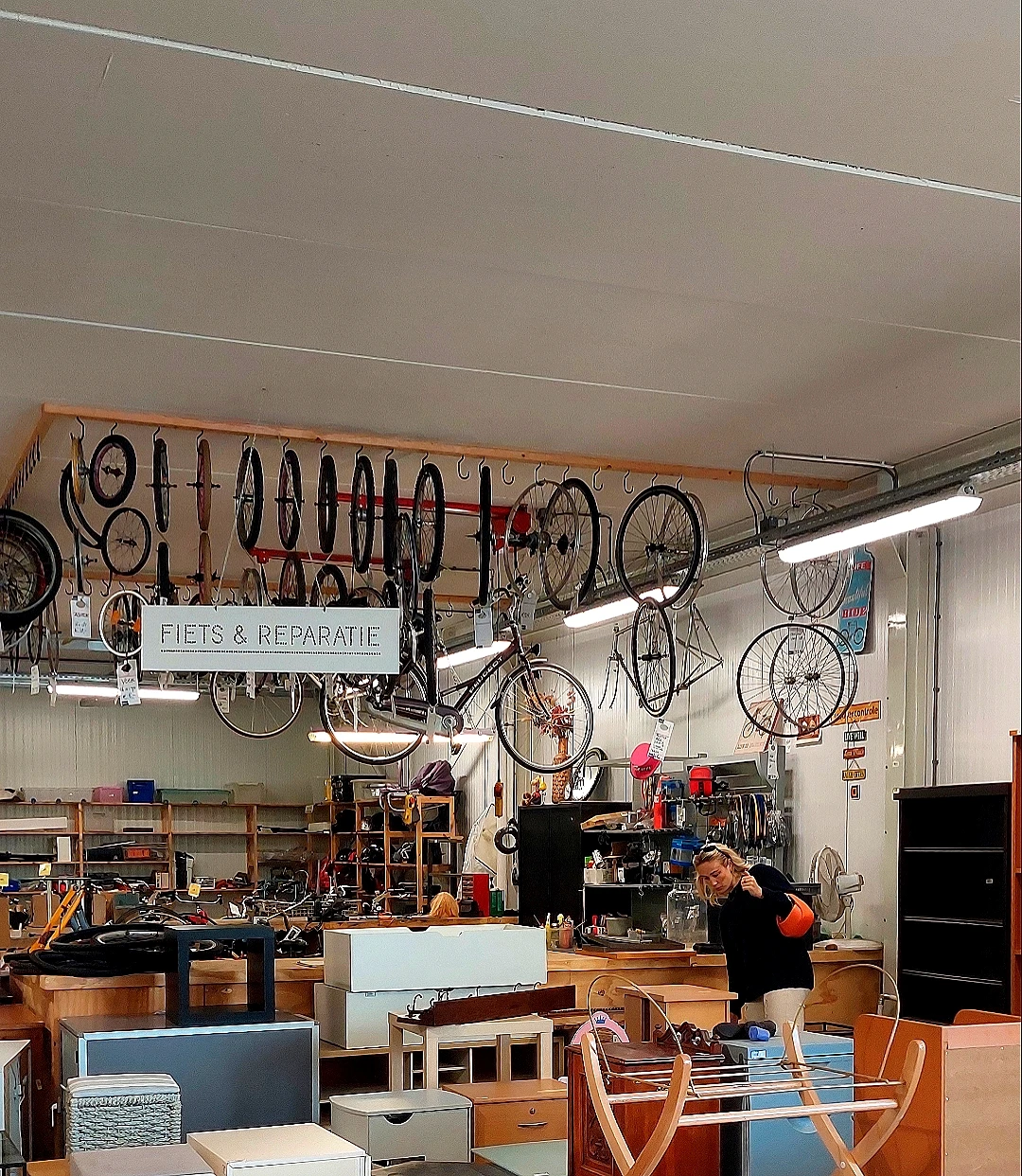 Werkplaats met fietsen aan wandrek en montage-werkstation, bord 'FIETS & REPARATIE' zichtbaar
