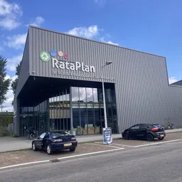Voorgevel van kringloopwinkel RataPlan met grote letters op donkergrijs pand, parkeerplaats met auto's ervoor en blauwe lucht