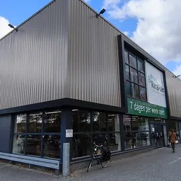 Voorgevel van kringloopwinkel met grijze golfplaten, grote ramen en groene reclamebord '7 dagen per week open'