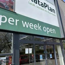 Groene gevel met tekst 'per week open' en ingang van kringloopwinkel met donkere deuren en ramen