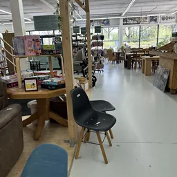 Winkelvloer met houten tafels, zwarte stoelen en stellingen met diverse spullen; grote ramen met uitzicht op groen