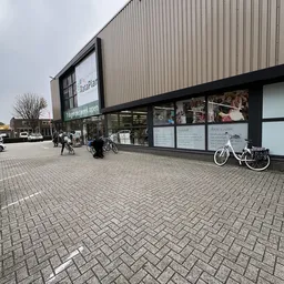 Gevel van kringloopwinkel RataPlan met etalageruiten, winkelnaam zichtbaar en fietsen ernaast op het plein