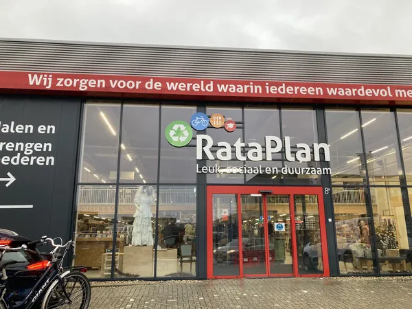 Gevel van kringloopwinkel RataPlan met rode deuren en grote ramen, naam duidelijk zichtbaar