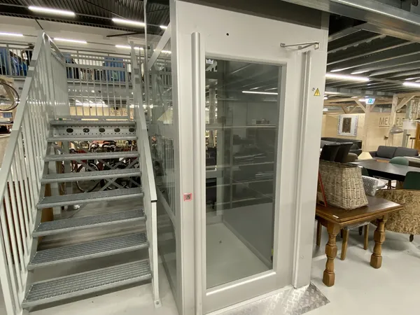 Witte showroom met glazen deuren en witte trap met metalen balustrade, op de achtergrond meubels en spullen uitgestald
