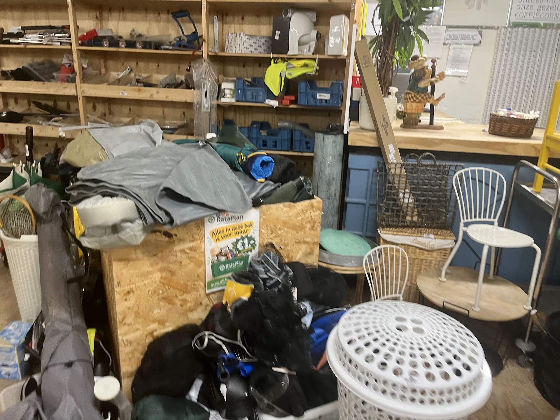 Rommelig interieur van kringloopwinkel met diverse spullen, dozen, stoelen en huishoudartikelen door elkaar