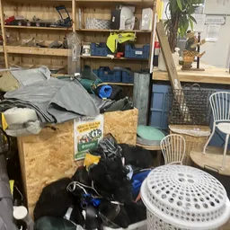 Rommelig interieur van kringloopwinkel met diverse spullen, dozen, stoelen en huishoudartikelen door elkaar