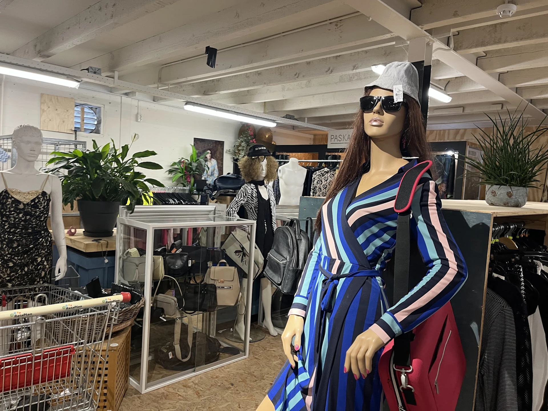 Etalagepop in blauwe gestreepte jurk met rode tas in kringloopwinkel, omgeven door rekken met kleding
