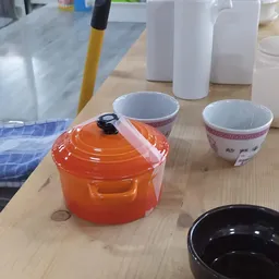 Oranje gesloten braadpan met zwarte knop op houten tafel naast twee decoratieve kopjes en witte containers