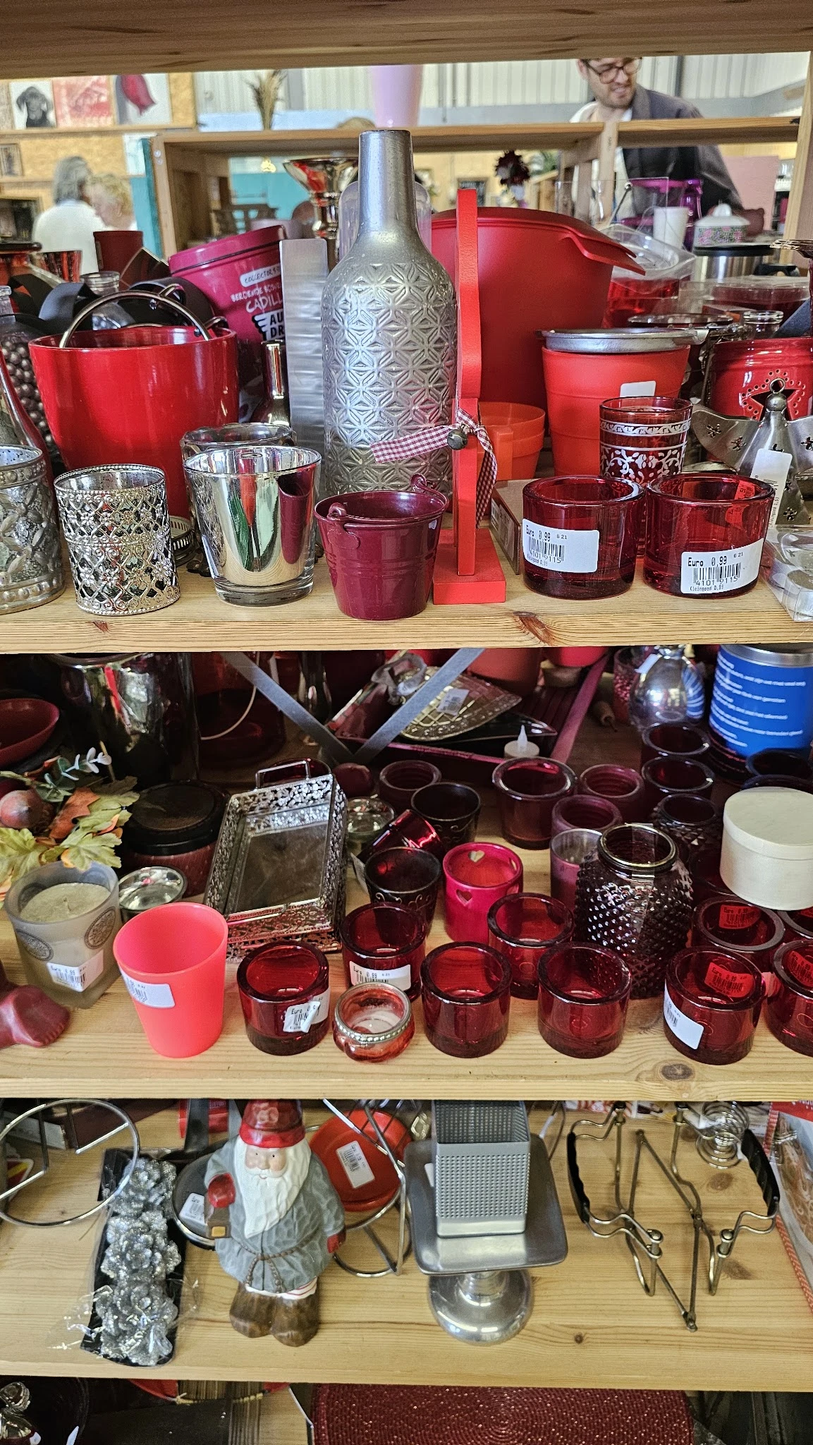 Uitgestalde kaarsen in verschillende kleuren en stijlen op planken, waaronder rode en roze kaarsen in glas