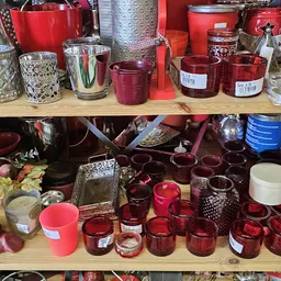Uitgestalde kaarsen in verschillende kleuren en stijlen op planken, waaronder rode en roze kaarsen in glas