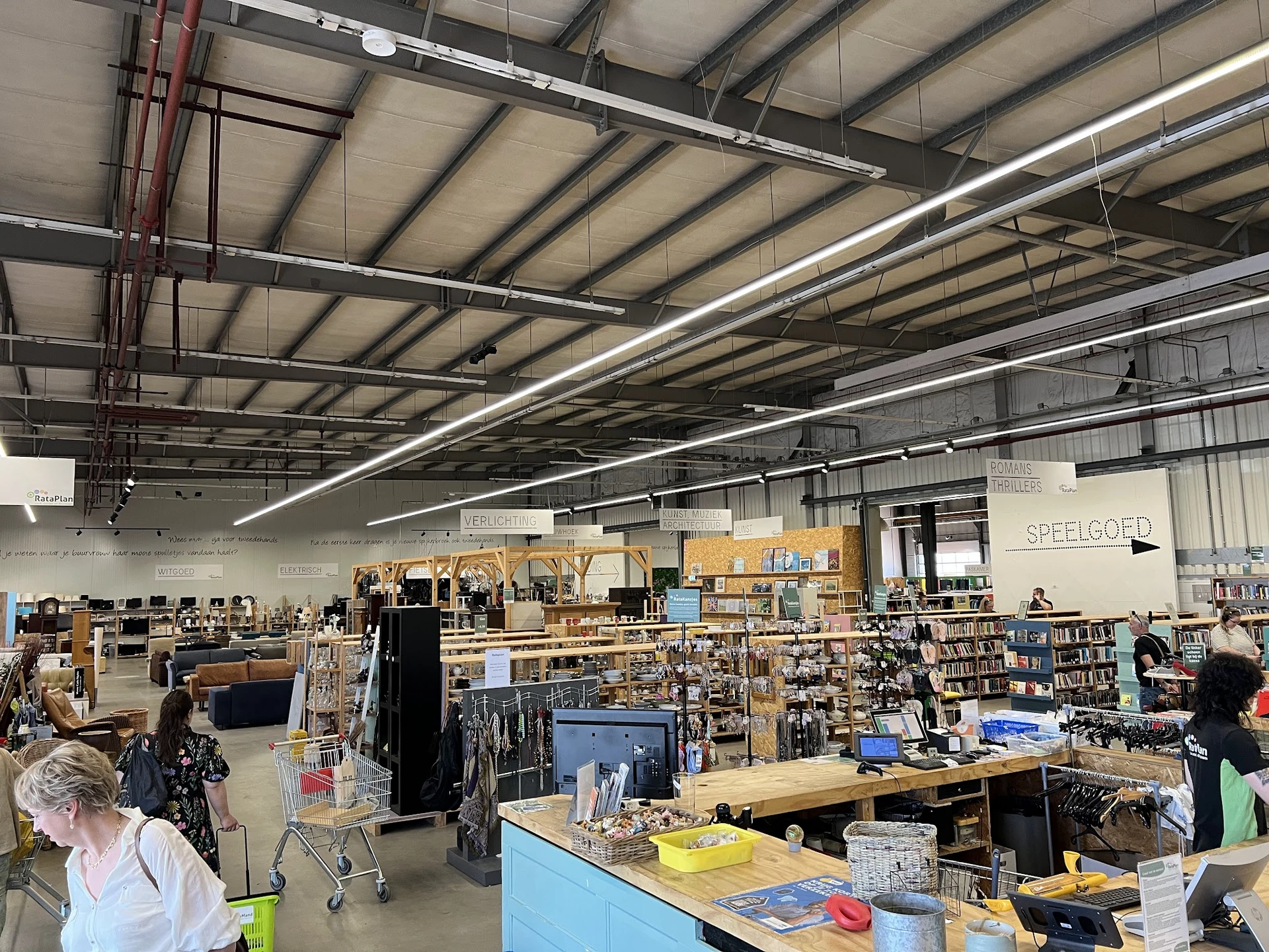 Ruim magazijn met houten stellingen vol spullen, tafels met artikelen en klanten aan het winkelen