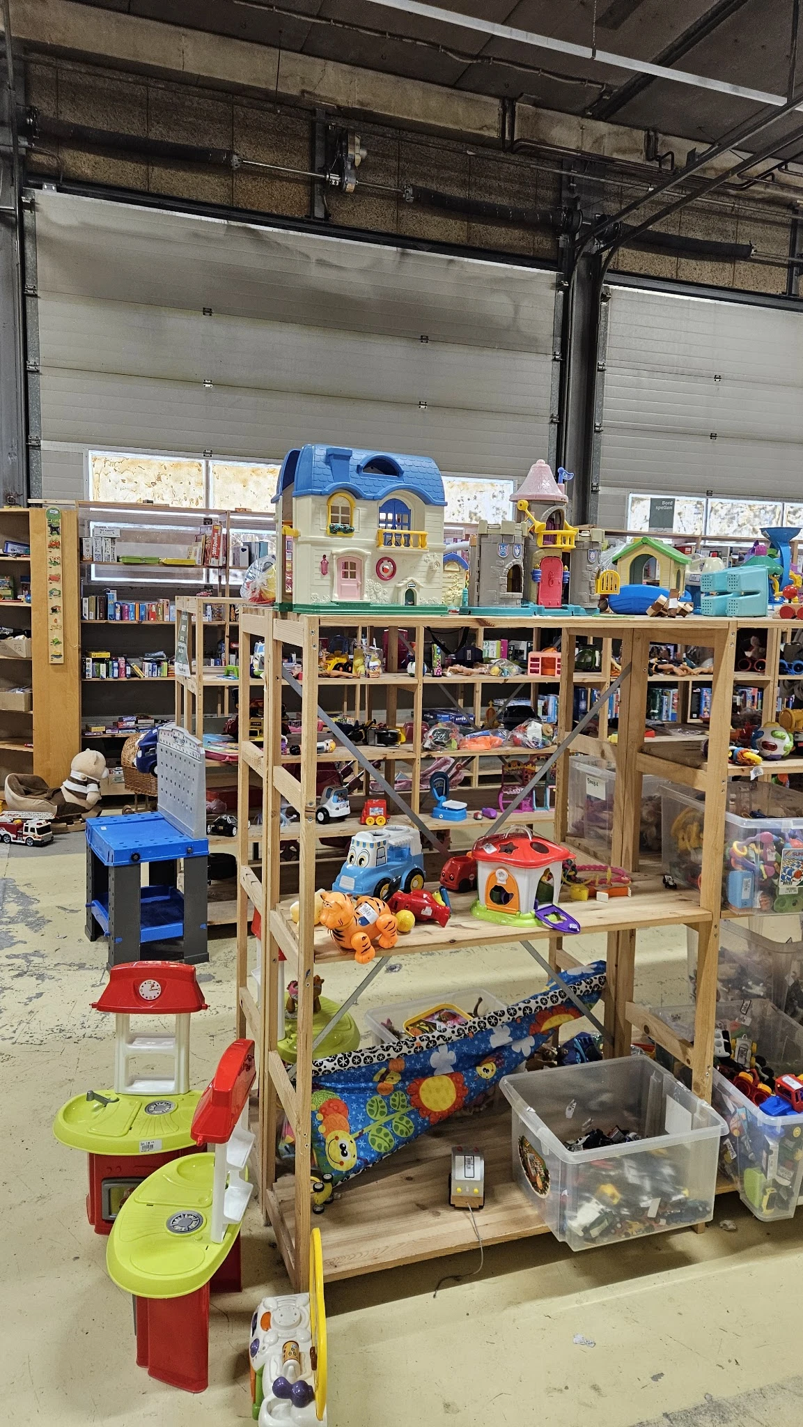 Speelgoedcollectie in kringloopwinkel: plastic huisjes, auto's, poppen en andere kinderspeelgoed op houten stellingen