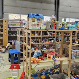 Speelgoedcollectie in kringloopwinkel: plastic huisjes, auto's, poppen en andere kinderspeelgoed op houten stellingen