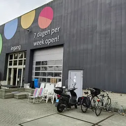 Grijze gevel van kringloopwinkel met gekleurde cirkels, ingang, raamdeuren en fietsen ervoor