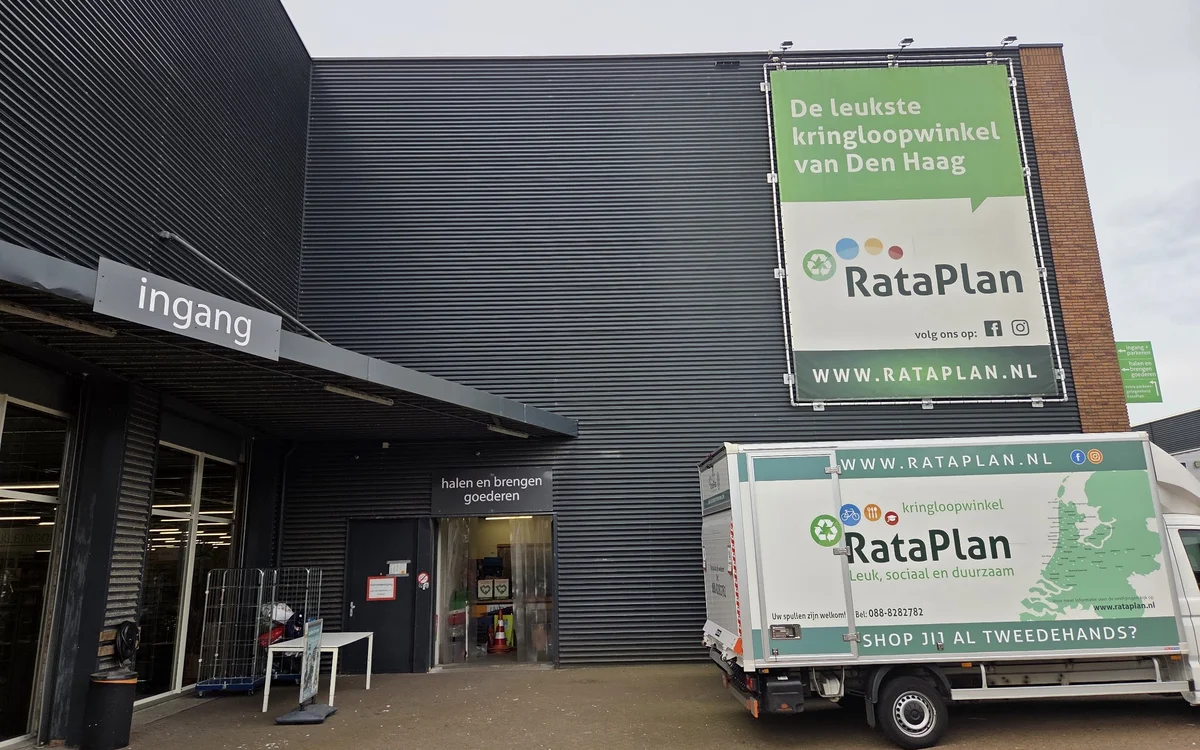 Voorkant kringloopwinkel met ingang, donkergrijze gevel en RataPlan borden. Bedrijfsbus geparkeerd ervoor.
