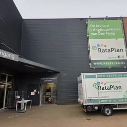 Voorkant kringloopwinkel met ingang, donkergrijze gevel en RataPlan borden. Bedrijfsbus geparkeerd ervoor.