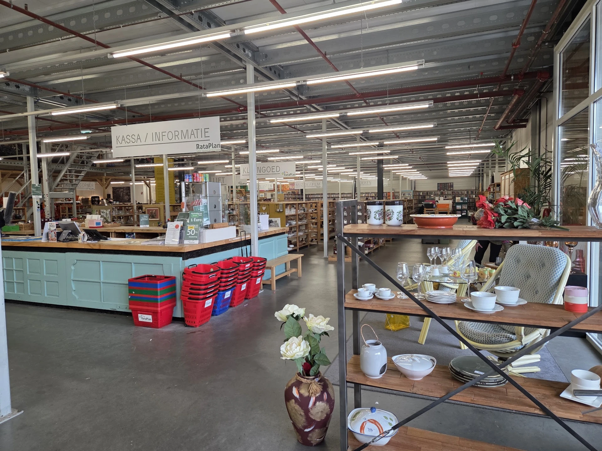 Overzicht van kringloopwinkel met kassabalie, stellingen met servies en huishoudwaar, rode kratten en wit beschilderde zuilen