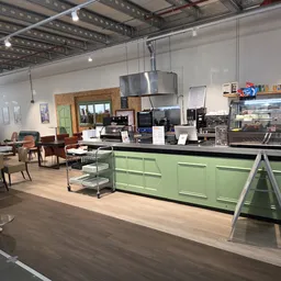 Winkelvloer met groen keukenblok, eettafels en diverse meubels en huishoudelijke artikelen.