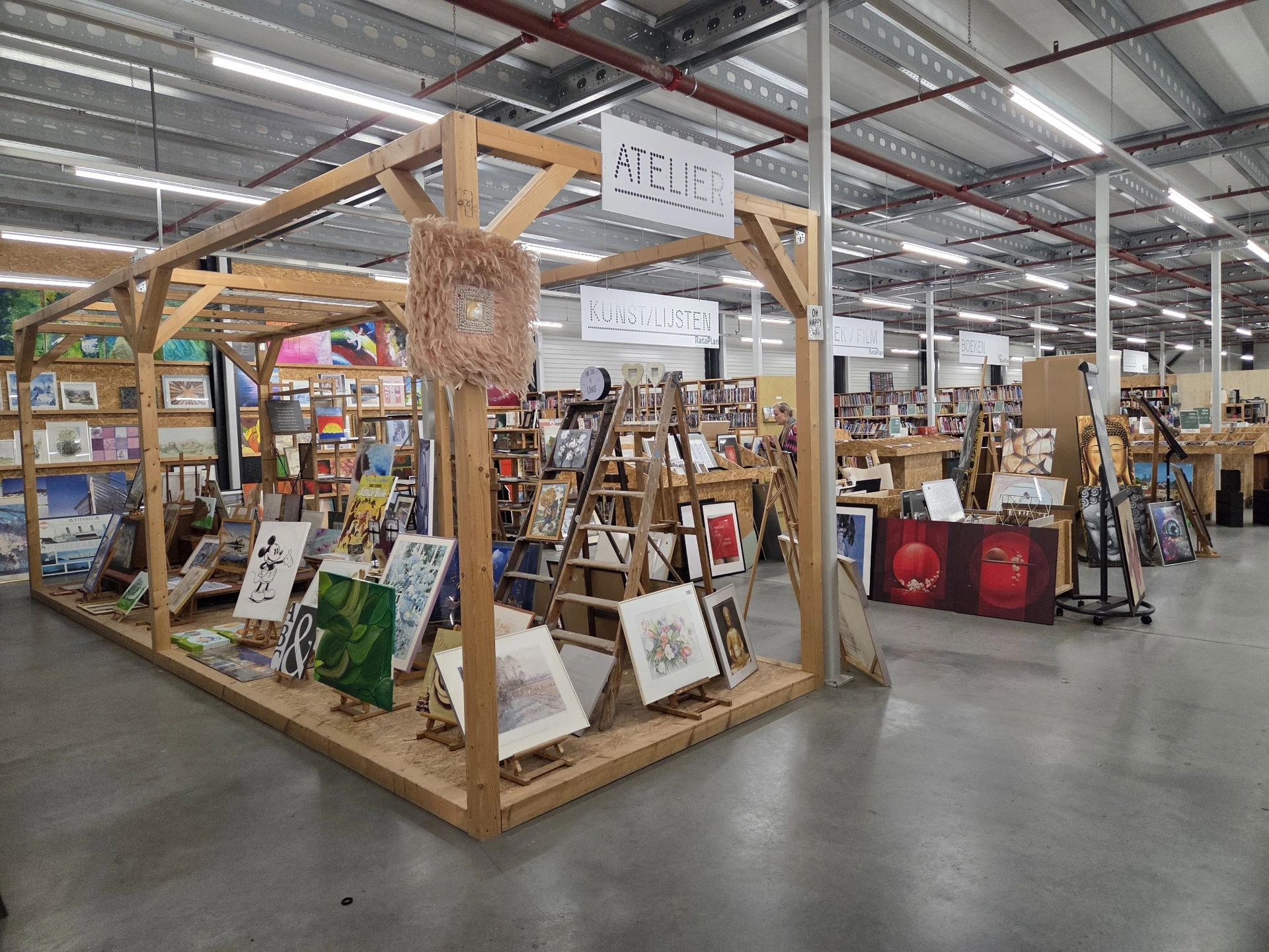 Winkelvloer met houten display-structuur vol kunstwerken, prints en schilderijen. Op de achtergrond meer stellingen met kunst