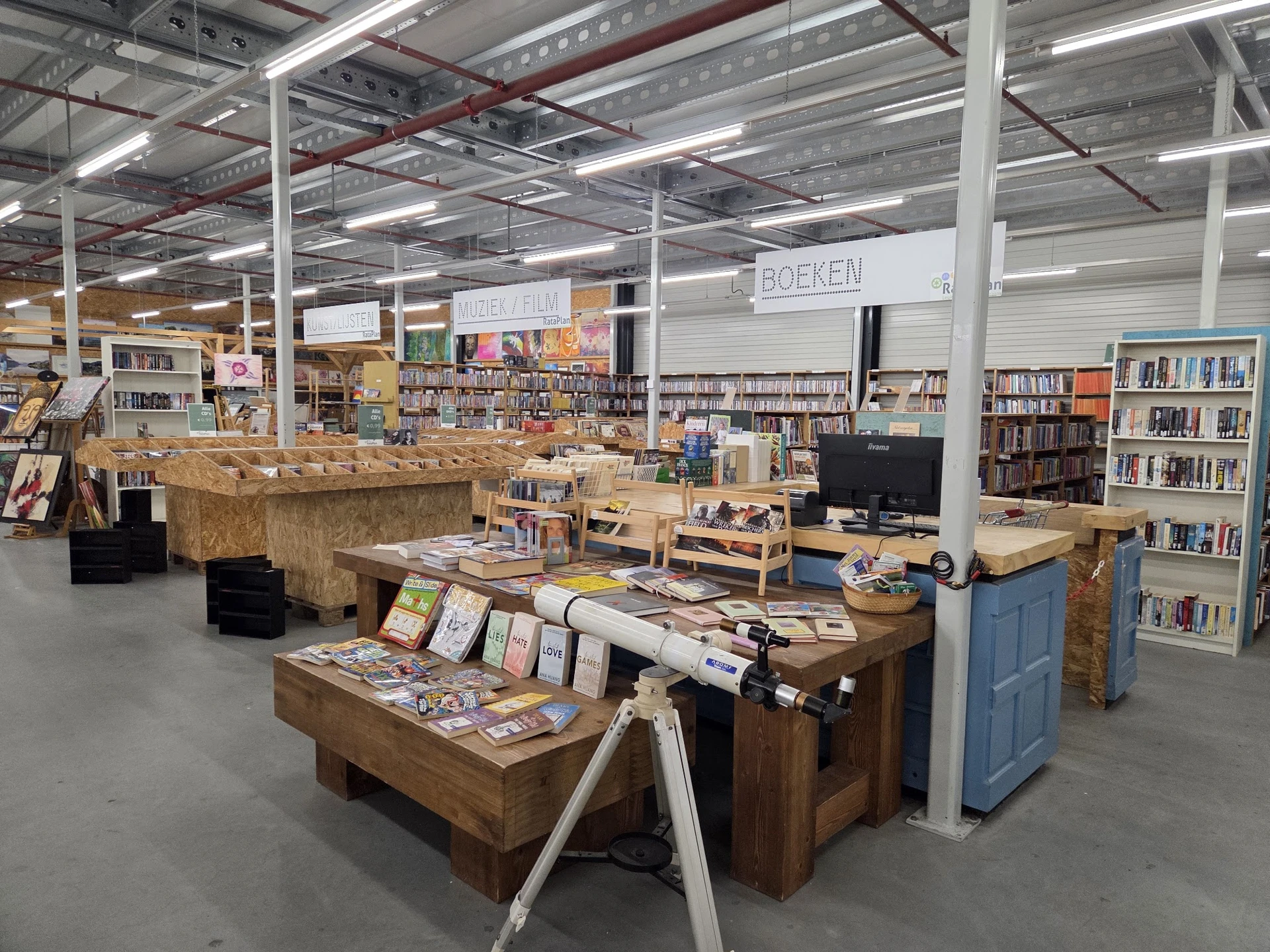 Winkelvloer met houten tafels vol boeken en andere spullen, stellingen met boeken op de achtergrond, industrieel plafond met 