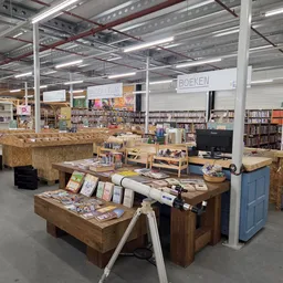 Winkelvloer met houten tafels vol boeken en andere spullen, stellingen met boeken op de achtergrond, industrieel plafond met 