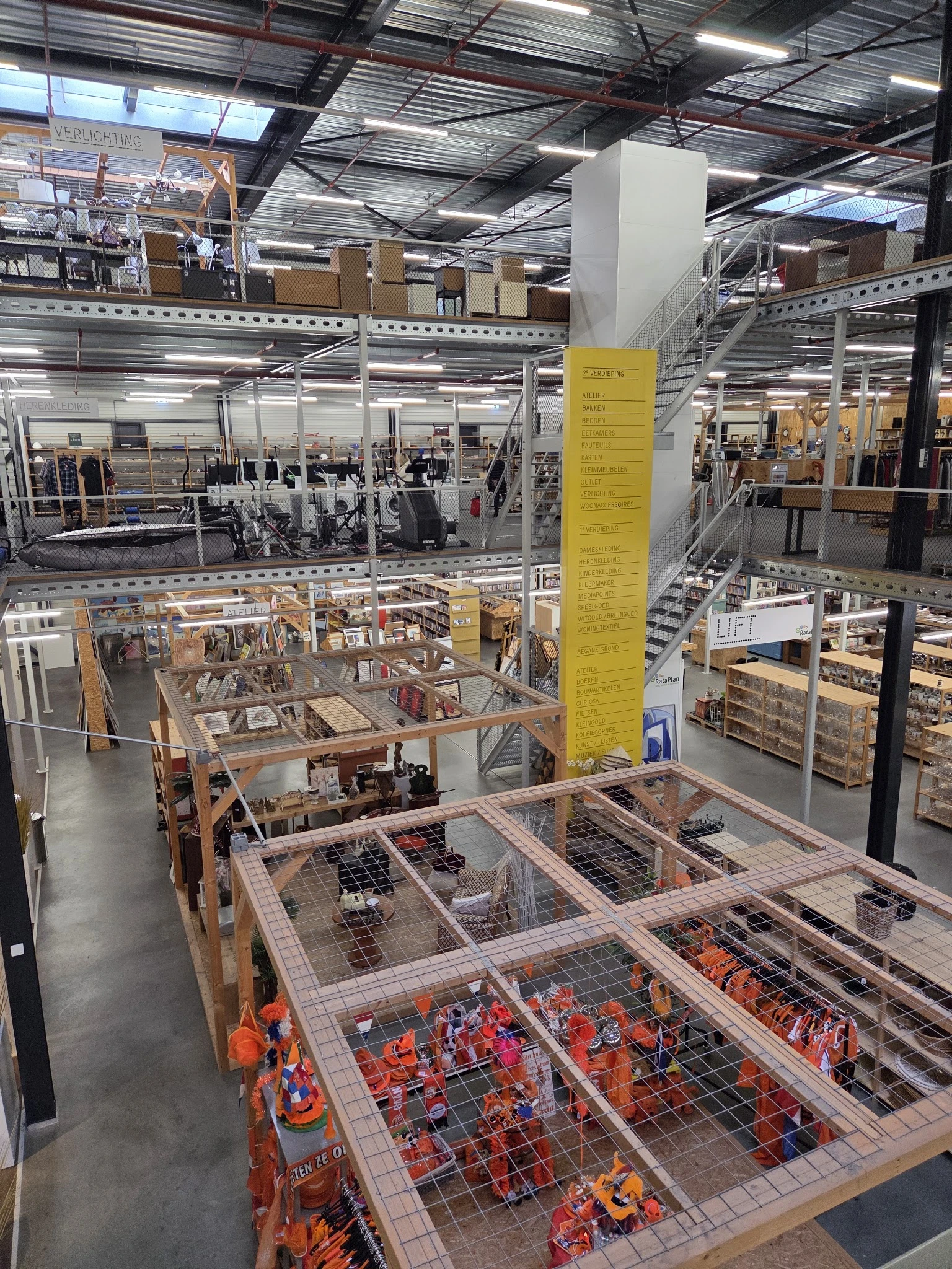 Meerdere verdiepingen van een kringloopwinkel met stellingen, opgeslagen spullen en een opvallende gele zuil in het midden