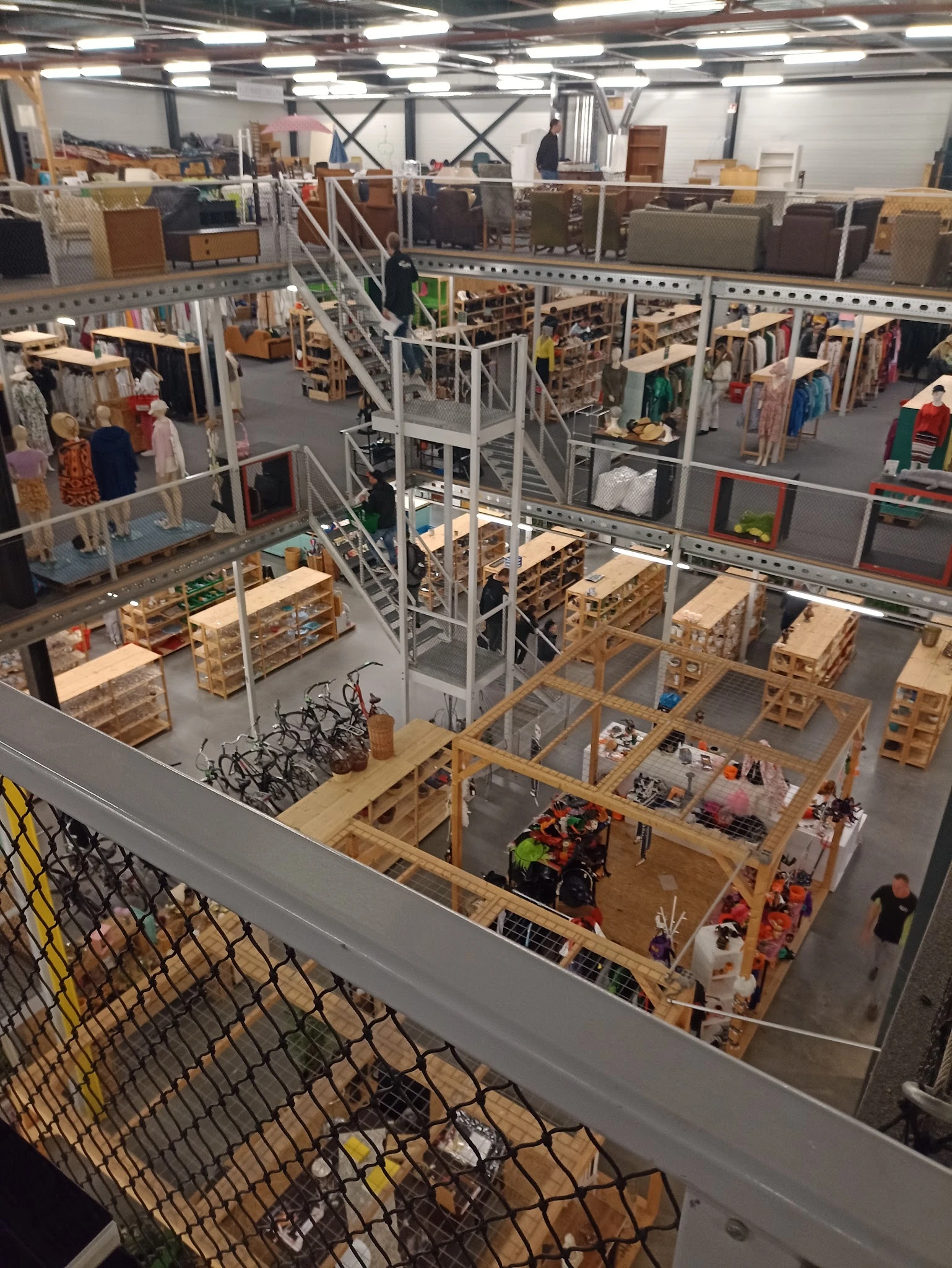 Overzicht van de kringloopwinkel met stellingen, kleding aan rekken, houten pallets en werknemers op de werkvloer