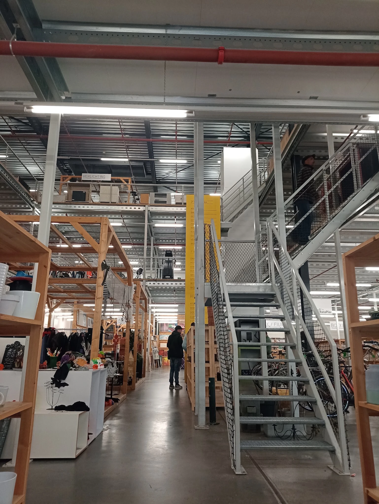 Ruim winkelinterieur met houten stellingen, metalen trappen en galerijen, winkeliers en bezoekers tussen de uitstalkanten