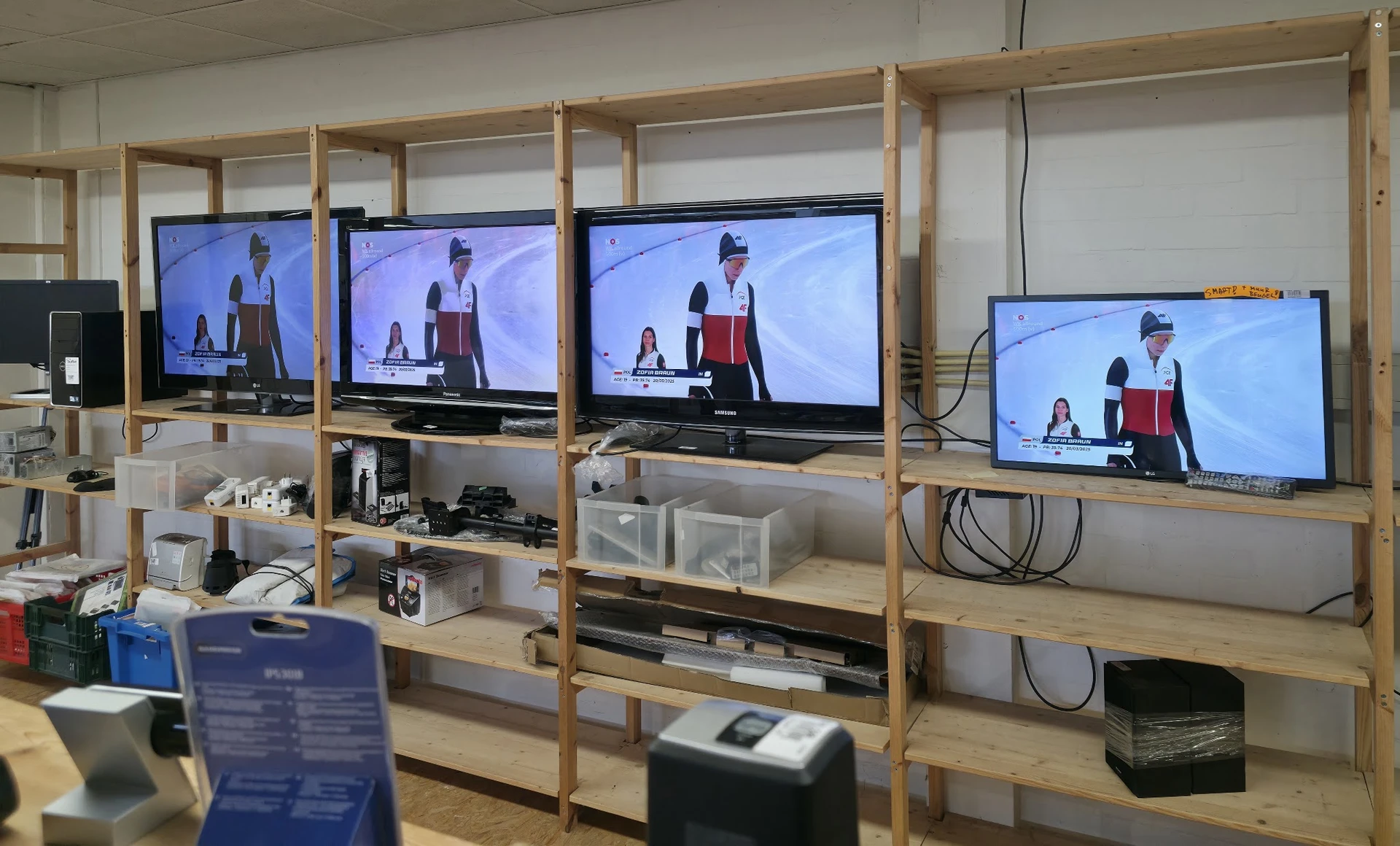 Houten stellingkasten met televisies waarop een presentator wordt getoond, met opbergdozen en spullen op de planken
