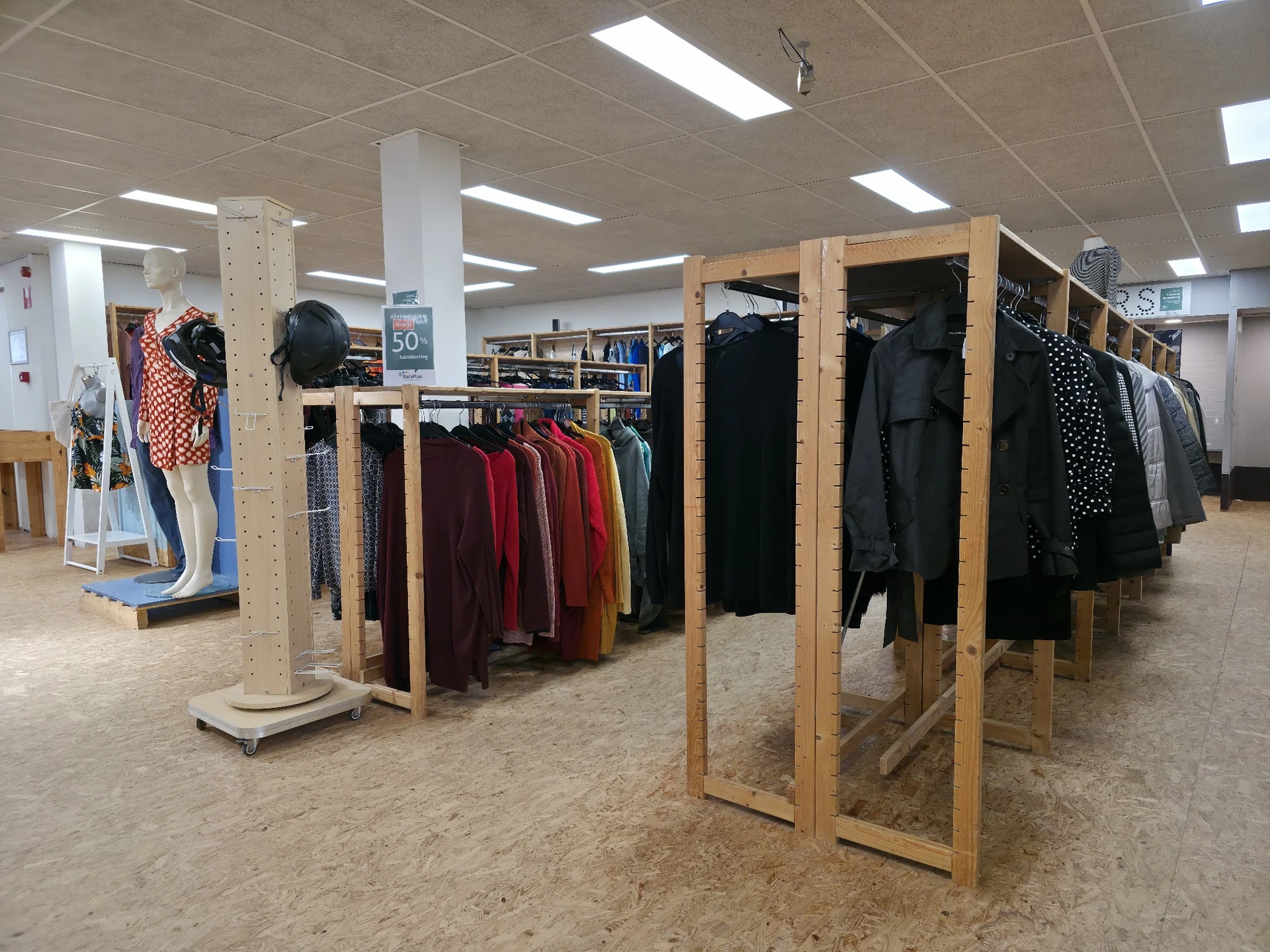 Winkelvloer met houten rekken vol tweedehands kleding, stellingen op de achtergrond en fluorescerende plafondverlichting