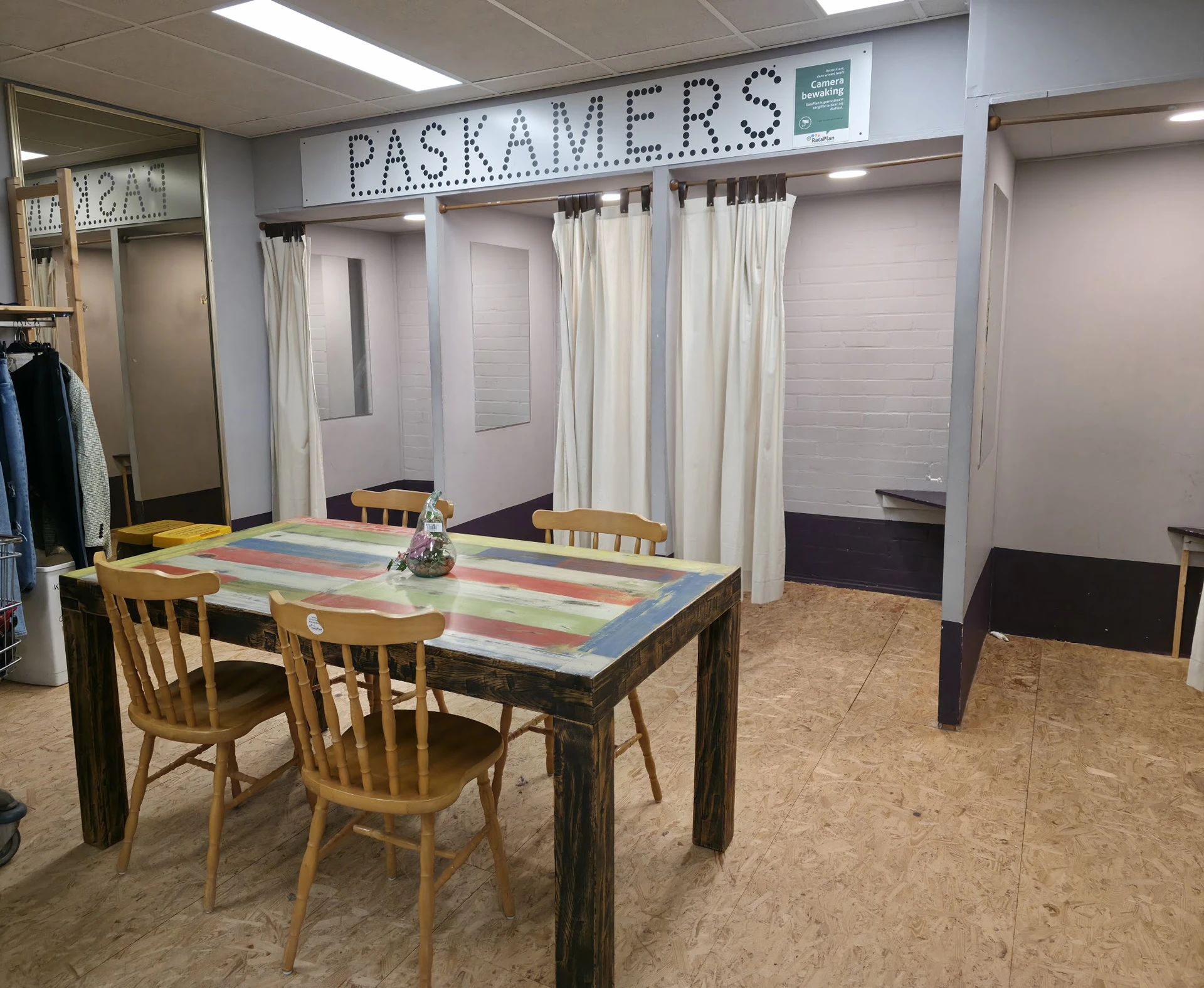 Winkelvloer met houten tafel, stoelen, gordijnen en winkelrekken met het opschrift 'PASKAMERS'